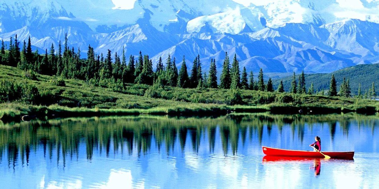 A person paddles a red canoe on a calm mountain lake surrounded by pine trees and snowy peaks.