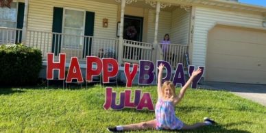 Girl in colorful dress doing a split in front of a birthday sign outside a house.