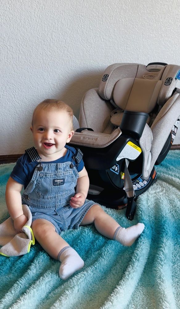 Smiling baby sitting on a blue blanket next to a beige car seat.