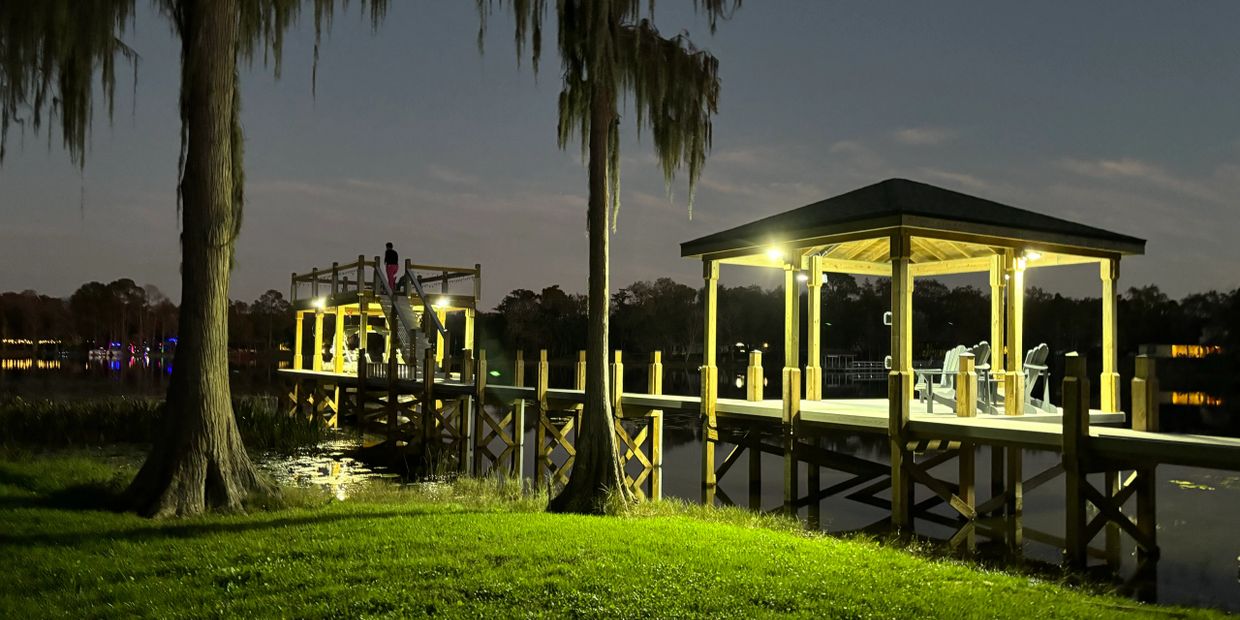 Lit wooden pier with a gazebo and people at dusk near calm water.