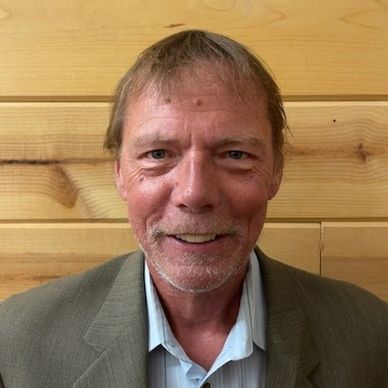 A middle-aged man in a suit jacket smiling against a wooden wall.