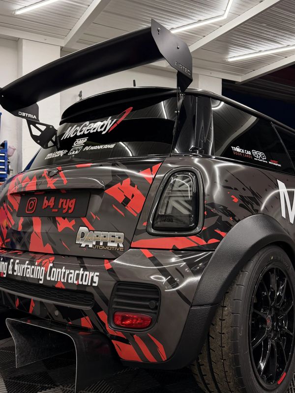 Customized Mini Cooper with racing decals and a large rear wing in a garage.