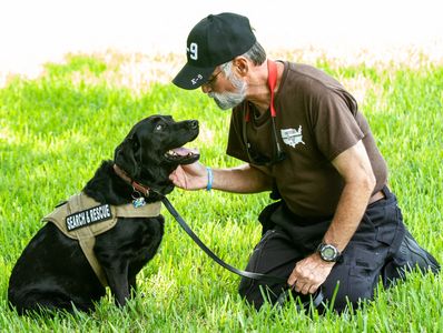 Man kneeling and petting a black search and rescue dog in a grassy area.