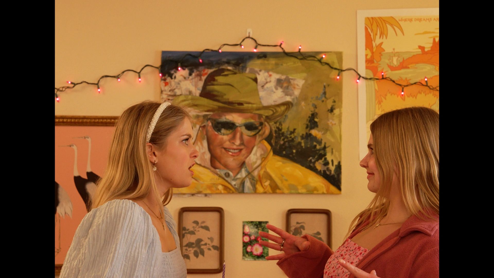 Two women facing each other, one surprised, with colorful artwork and string lights in the background.