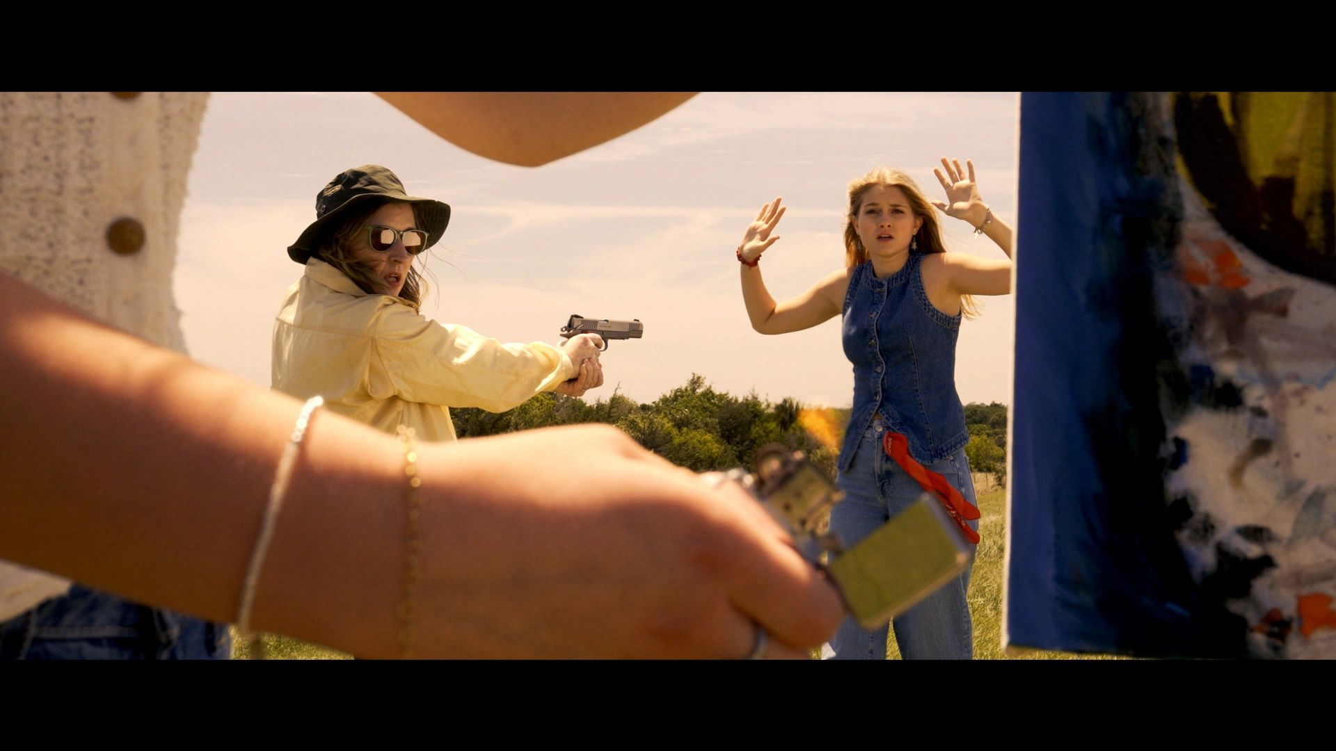 Woman in hat aiming gun at another woman with hands raised outdoors.