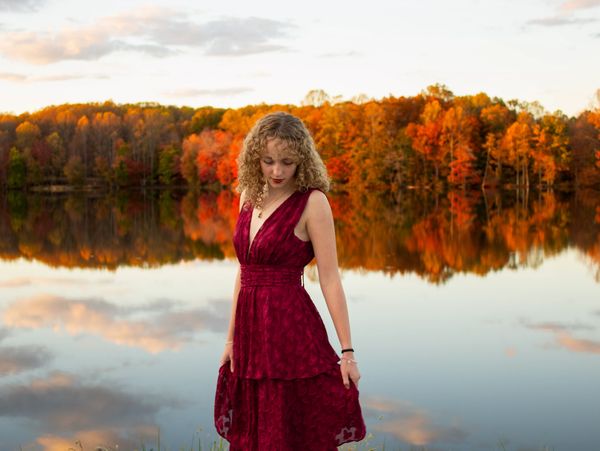 A woman in a red dress stands by a lake with autumn trees reflecting in the water.