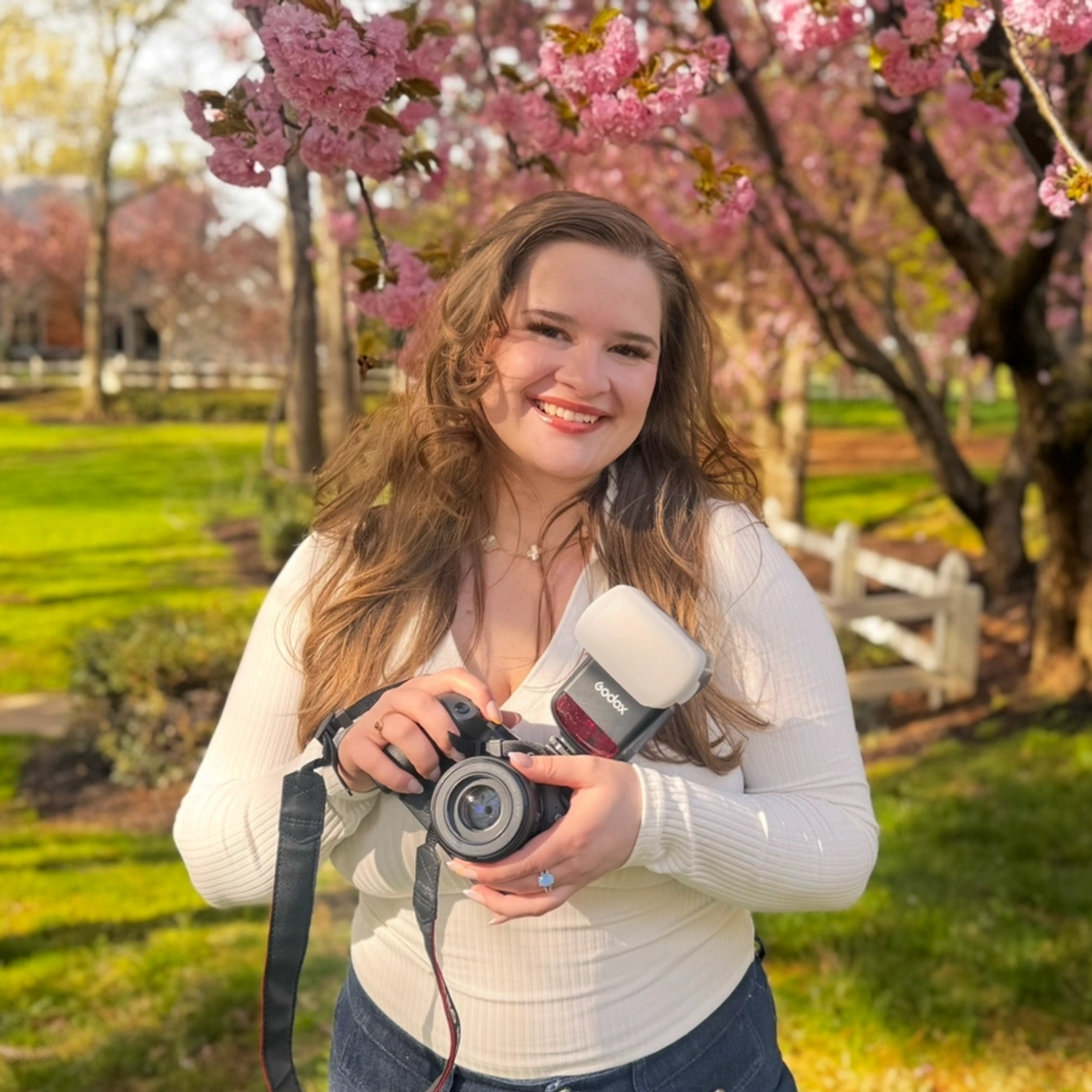 Smiling woman holding a camera among blooming cherry blossoms.
