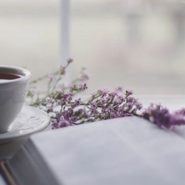 A cup of tea, purple flowers, and an open book on a table.