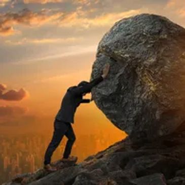 Person pushing a large rock uphill at sunset.