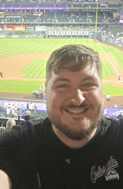 Man smiling at a baseball stadium wearing a Colorado Rockies shirt.