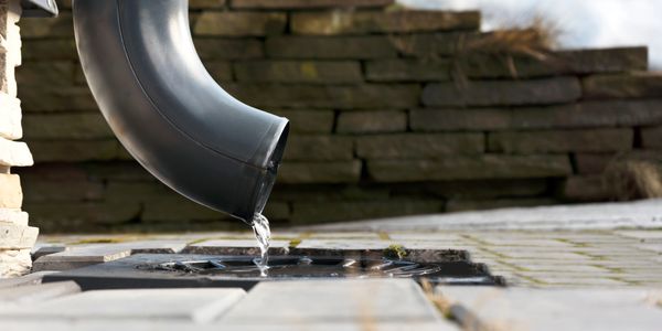 Water flows from a black pipe onto a wet ground surface outdoors.