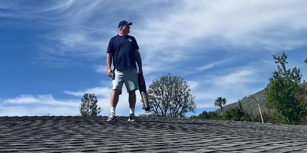 Man stands on roof holding a leaf blower under a partly cloudy sky.