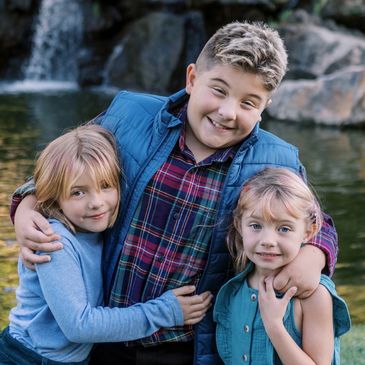 A big brother hugging his two younger sisters while all three children are smiling.