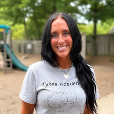 Smiling woman with long black hair wearing a Tykes Academy shirt outdoors.
