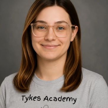 Smiling woman with glasses wearing a Tykes Academy shirt against a gray backdrop.