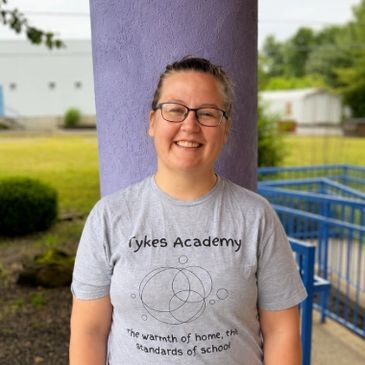 Smiling woman wearing a Sykes Academy t-shirt standing outdoors by a purple pillar.