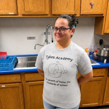 Woman smiling in a kitchen wearing a Tykes Academy shirt.