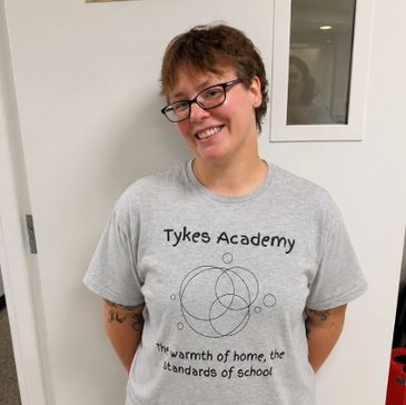 Person smiling wearing a Tykes Academy shirt with a welcoming message.