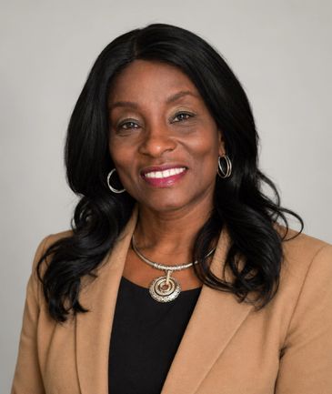 Smiling woman with black hair wearing a tan blazer and silver jewelry.