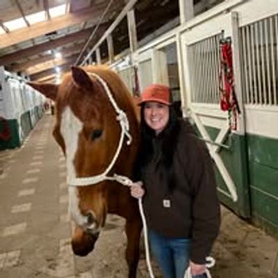 Woman standing beside a brown horse in a stable, holding its rope.