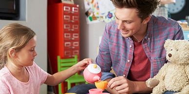 A young girl and man enjoy a tea party with a teddy bear.