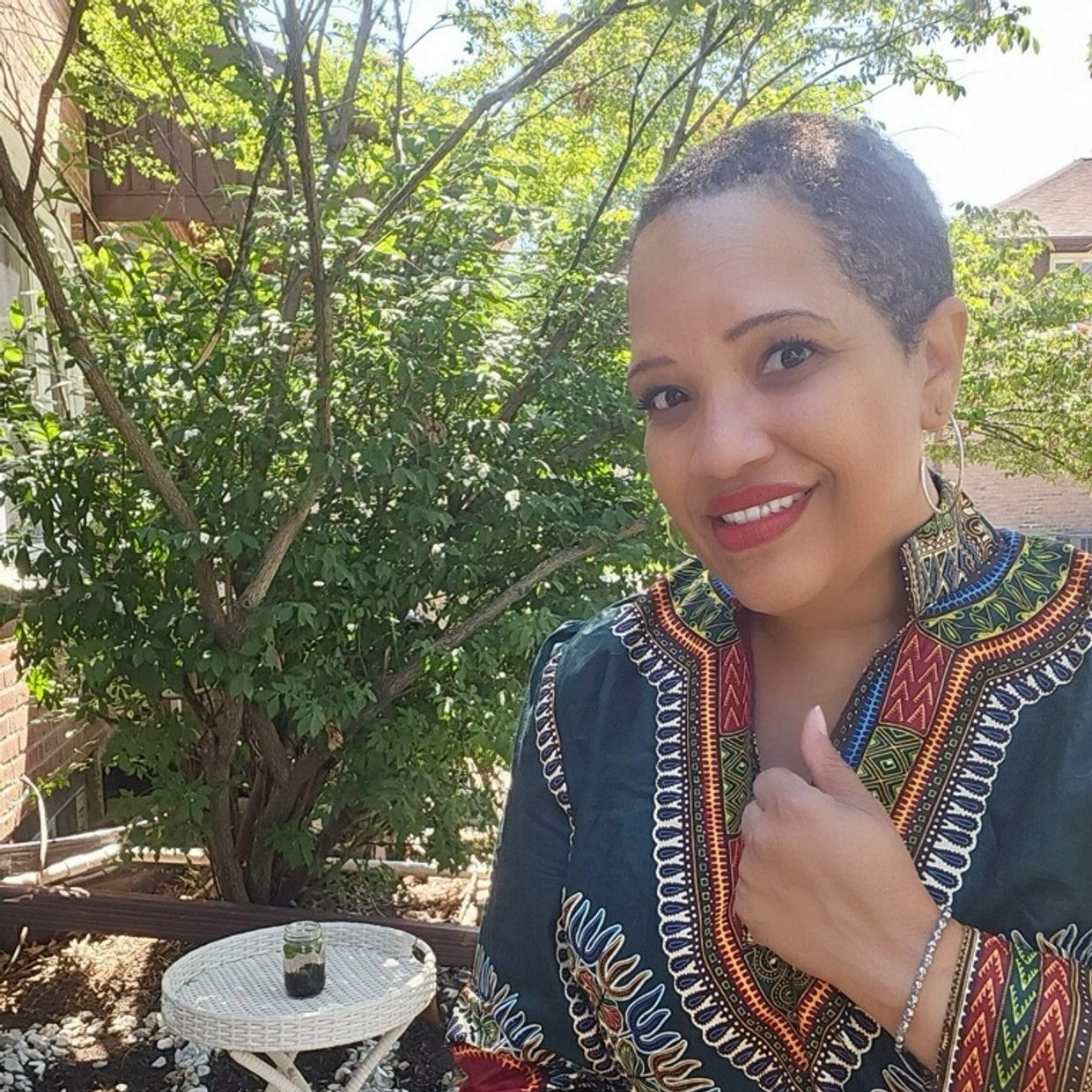 Smiling woman outdoors wearing a colorful patterned dress, sitting by a small white table.