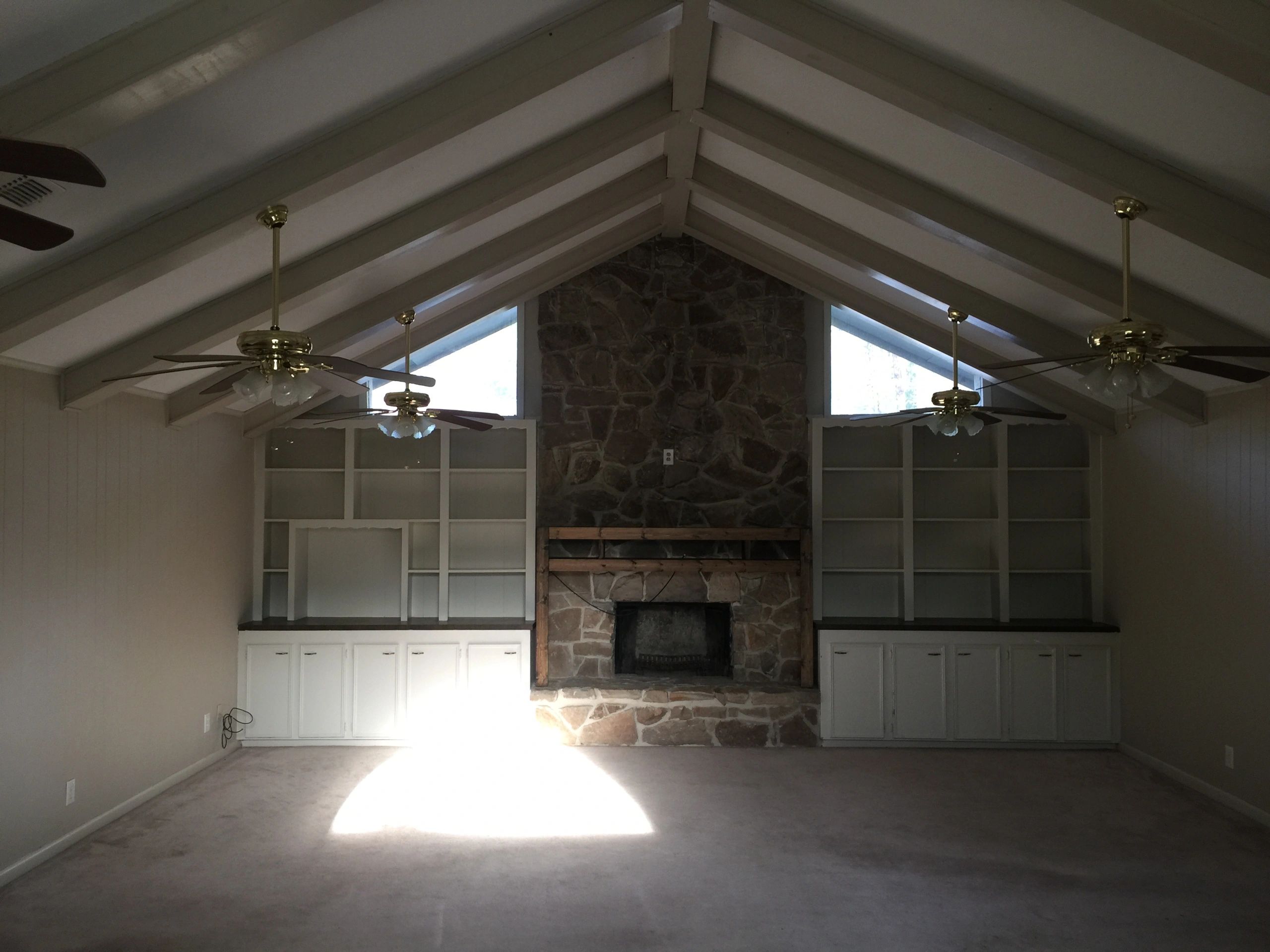 Empty living room with vaulted ceiling, stone fireplace, ceiling fans, and built-in shelves.