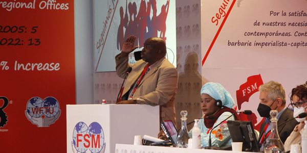 A man speaks at a podium during an FSM conference with attendees listening.