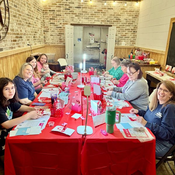 A group of women and a baby crafting at a long red table in a cozy room.