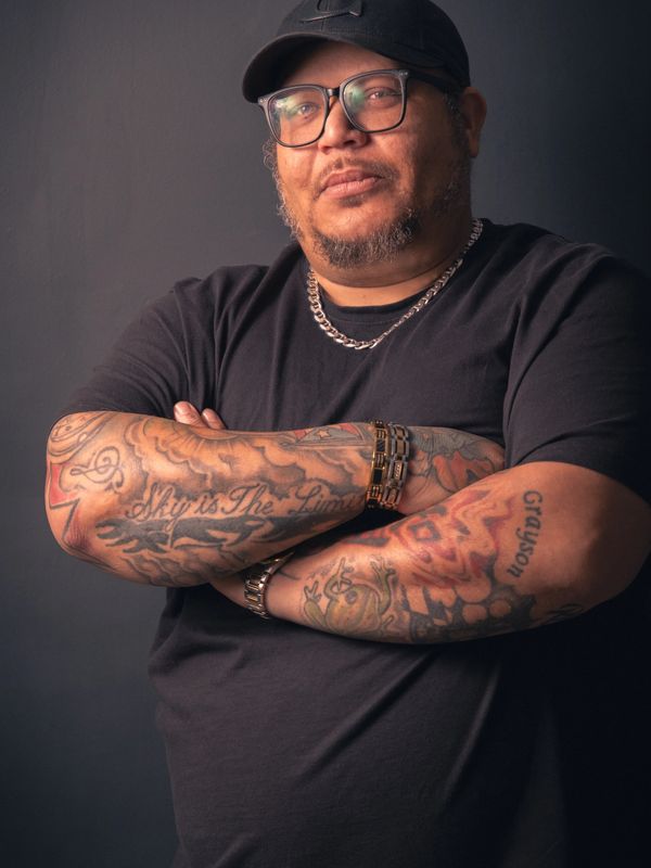 Man with tattoos on arms wearing black shirt and cap.