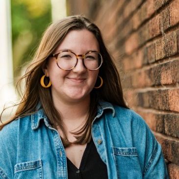 Young woman smiling by a brick wall wearing glasses and denim jacket.