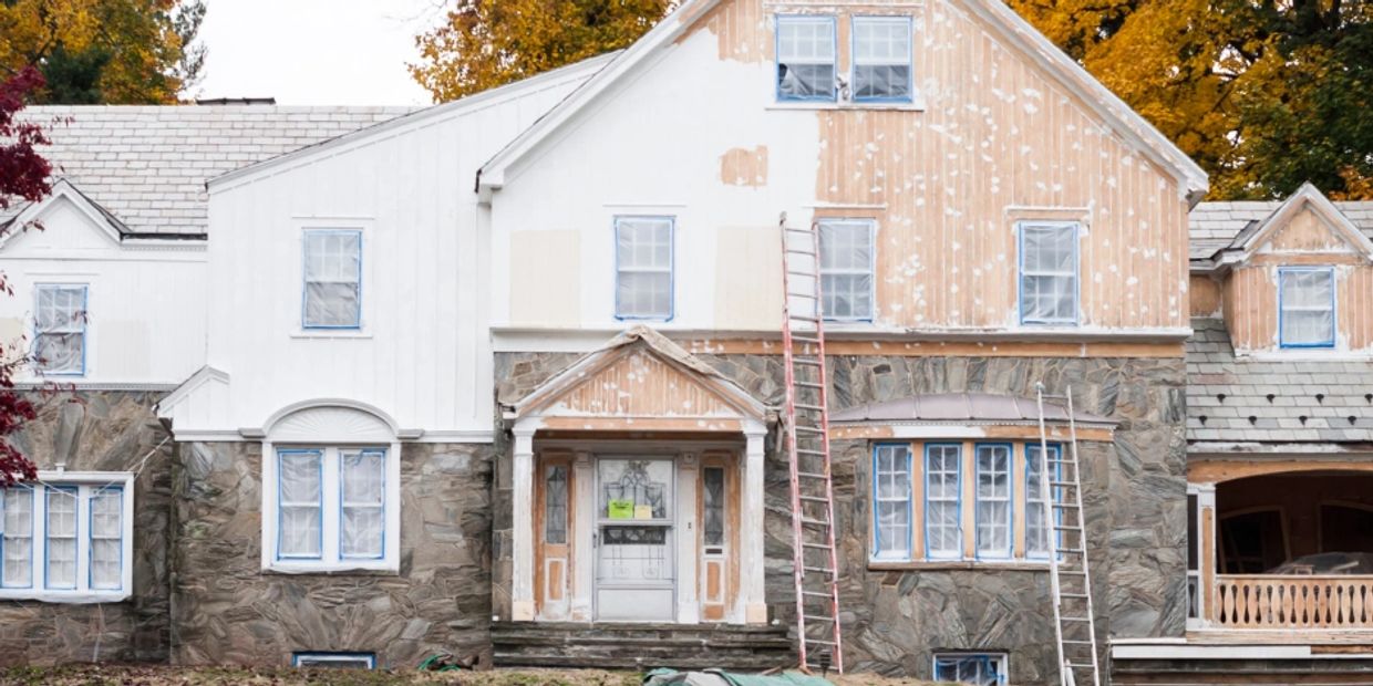A large house undergoing exterior renovations with ladders and paint preparation.