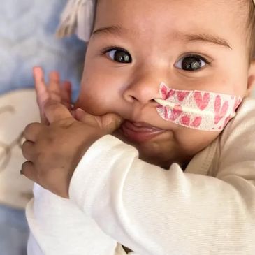 A baby with a heart-patterned medical tape on the cheek sucking thumb.