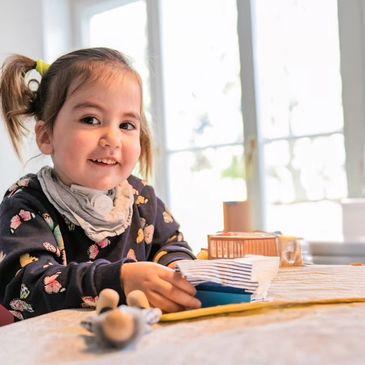 Smiling young girl playing with toys at a table by a window.