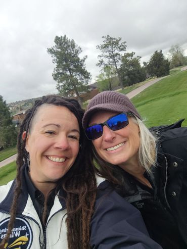 Two women smiling outdoors on a cloudy day with trees and grass in the background.