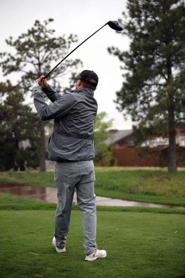 Golfer in gray jacket and cap swings club on a green golf course.