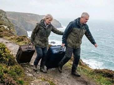 Two people hiking with suitcases along a coastal cliff path.