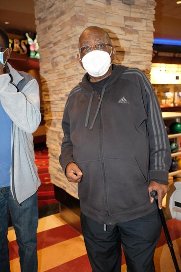 Man wearing face mask and dark Adidas jacket standing with cane indoors.
