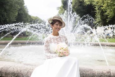 Bride in white dress holding bouquet by a fountain.