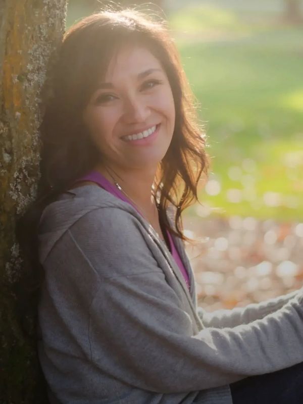 A woman smiling warmly while leaning against a tree outdoors.