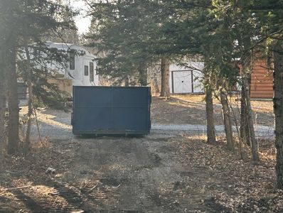 A large blue dumpster on a dirt driveway surrounded by trees.