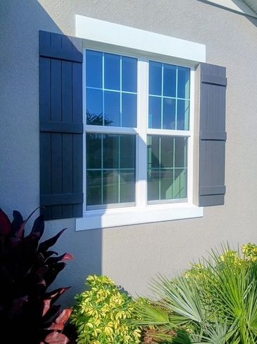 A bright window with dark shutters on a beige wall, surrounded by green and red plants.