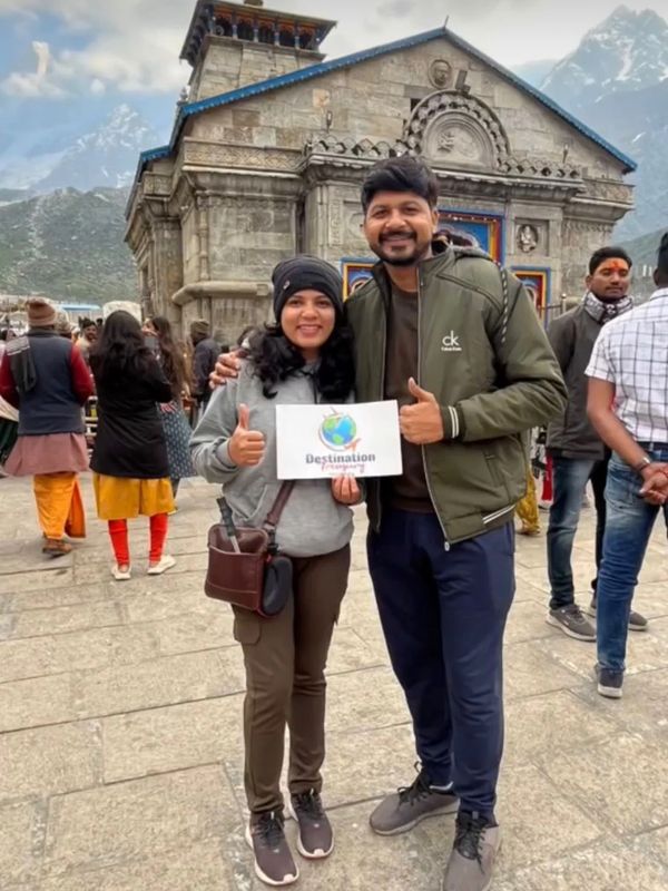 A smiling couple poses with a destination sign in front of a historic stone temple.