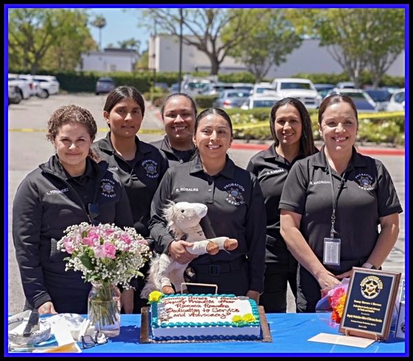 Six smiling women in black uniforms behind a decorated table with a cake and flowers outdoors.
