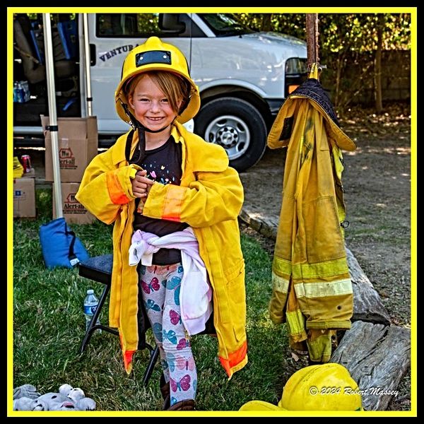 A young child smiling, dressed in oversized firefighter gear outdoors.