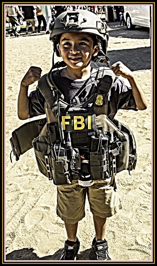 Young boy flexing muscles in oversized FBI tactical gear and helmet outdoors.