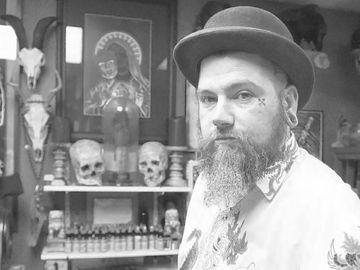 Bearded tattoo artist in a studio with skulls and artwork.