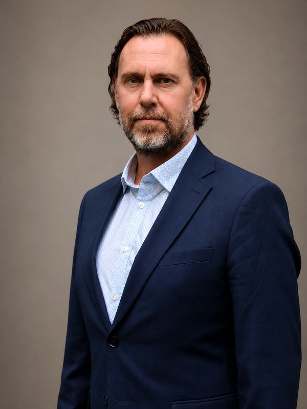 Confident middle-aged man in a navy suit and light blue shirt posing against a neutral background.