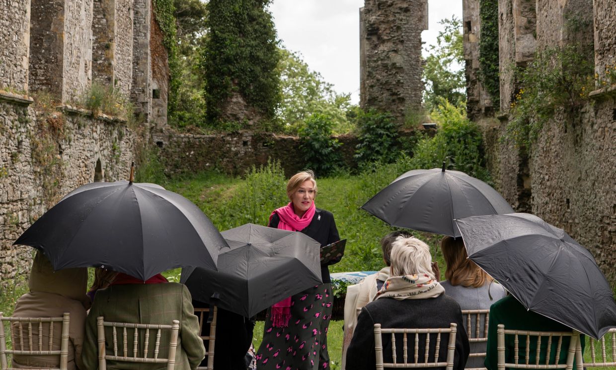 Woman speaking to an audience with umbrellas in ruins.