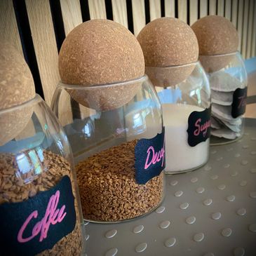 Glass jars with cork lids holding coffee, decaf, sugar, and salt on a shelf.